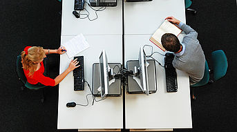 Das Bild zeigt einige Computerarbeitsplätze von oben. An einem Computer sitzt eine Studentin. An einem anderen Computer sitzt ein Student.