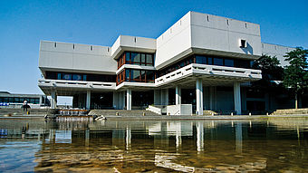 Das Bild zeigt die Uni-Bibliothek von außen. Davor sieht man den Teich, der auf dem Gelände von der Universität ist.